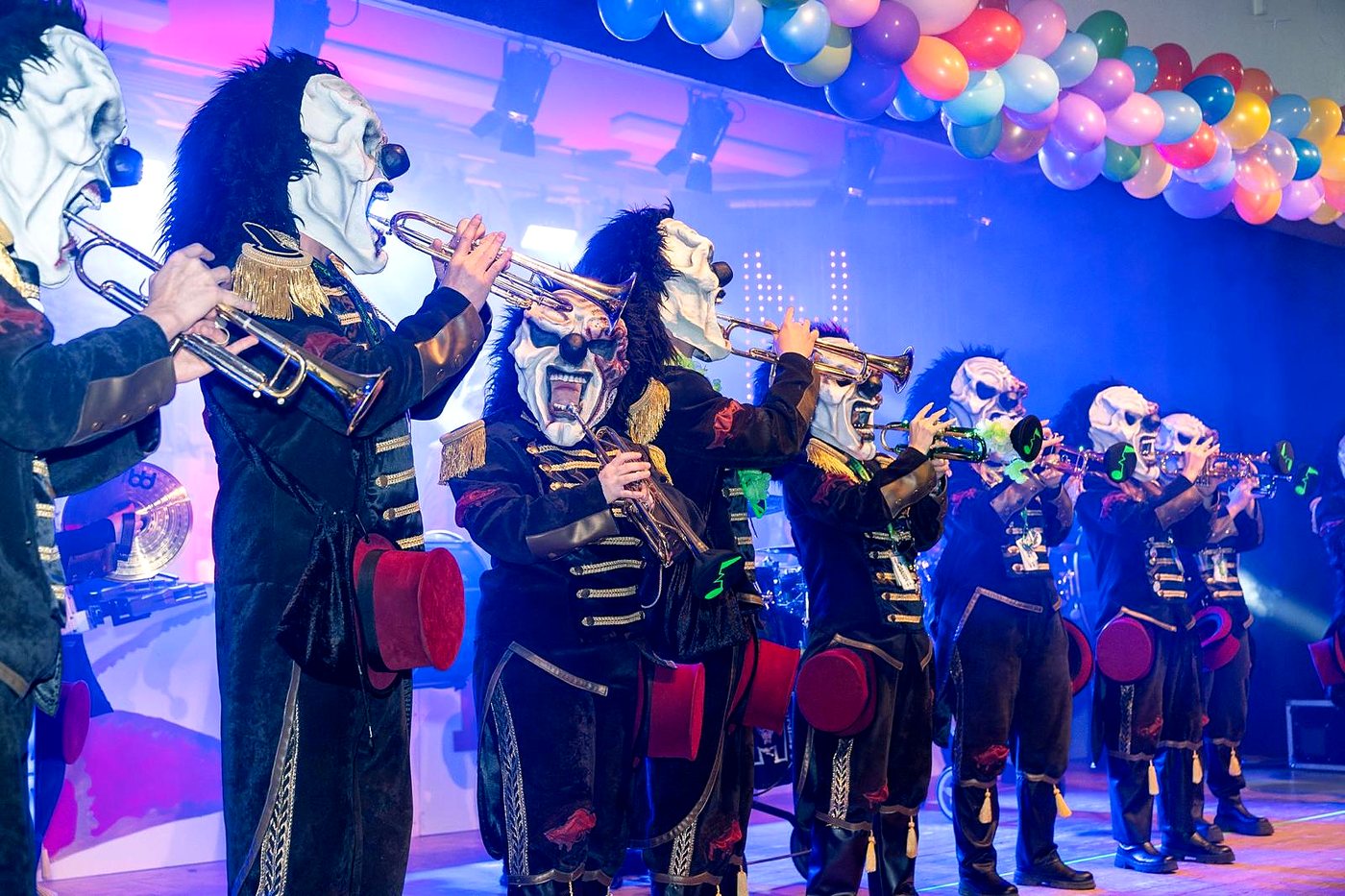 Die Lärmer auf der Bühne: Trompeten, Horror-Clown-Masken, schwarze Uniformjacken mit Goldborten und rote Zylinder am Gürtel, darüber bunte Luftballons
