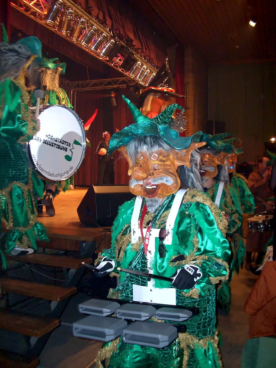 Lärmer in grünem Kostüm mit Maske und spitzem Hut am Percussion-Set, daneben eine Trommel mit Lärmer-Logo